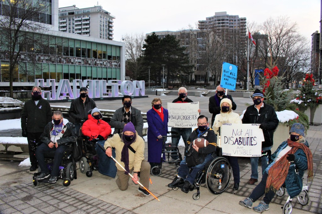 This photo was taken on December 3rd, 2020. It was taken in front of City Hall and has several members of the ACPD posing for the photo with the Hamilton sign in the background. There are thirteen people arrayed in a semicircle. They are all masked and dressed for cold weather. Some are holding signs in support of person with invisible disabilities.